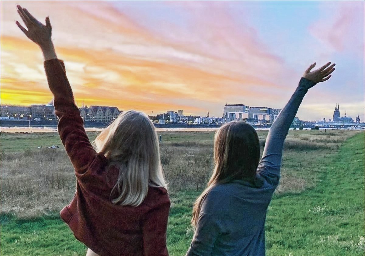 Zwei Menschen blicken bei warmem Abendlicht auf die Kölner Skyline und heben zuversichtlich die Arme